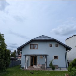 Grzegorz Zabrzeński EKOPRO - Błękitny dom jednorodzinny z szarym dachem i brązowymi ramami okiennymi, otoczony zielonym ogrodem pod pochmurnym niebem. Widoczna pergola z białymi zasłonami.