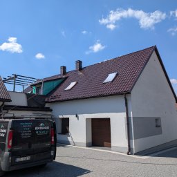 Grzegorz Zabrzeński EKOPRO - Dom po renowacji z nowym, brązowym dachem i jasną elewacją. Na podjeździe stoi firmowy van z reklamą usług malowania i mycia dachów oraz elewacji.