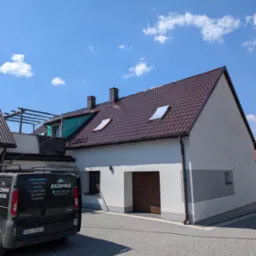 Dom po renowacji z nowym, brązowym dachem i jasną elewacją. Na podjeździe stoi firmowy van z reklamą usług malowania i mycia dachów oraz elewacji.