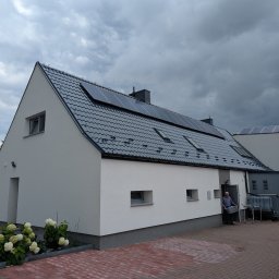 Grzegorz Zabrzeński EKOPRO - Biały dom z ciemnoszarą dachówką i panelami solarnymi. Widoczny fragment kostki brukowej i osoba w drzwiach. Pochmurne niebo w tle.