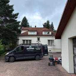 Grzegorz Zabrzeński EKOPRO - Pracownik na dachu domu z czerwonej dachówki, obok zaparkowany czarny bus firmy EkoPro Malowanie Dachów i sprzęt do mycia pod ciśnieniem. Pochmurne niebo.