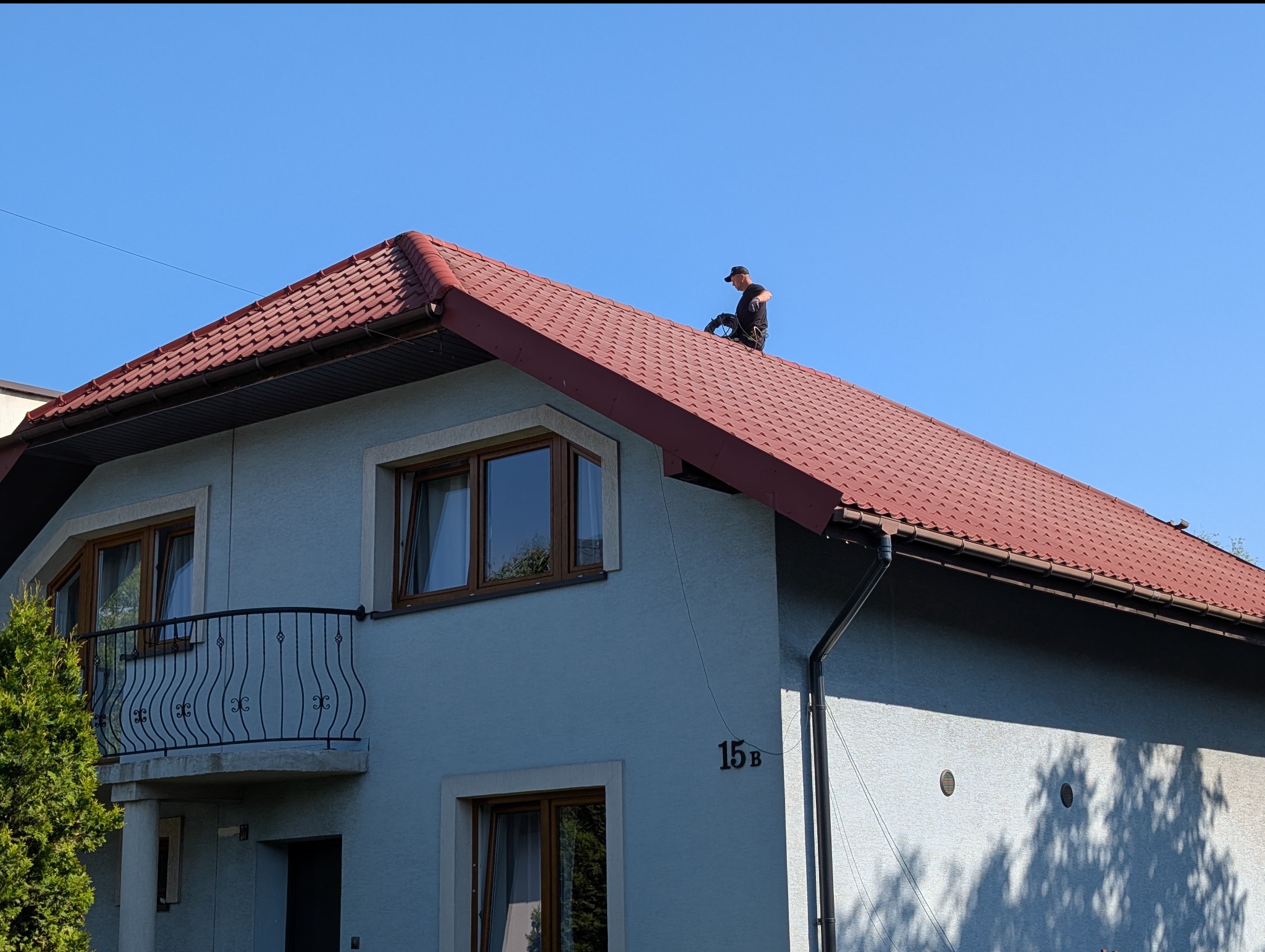 Pracownik w czarnym stroju na czerwonym dachu dwupiętrowego domu z jasną elewacją. Widok z dołu na tle błękitnego nieba, cień drzewa na ścianie.
