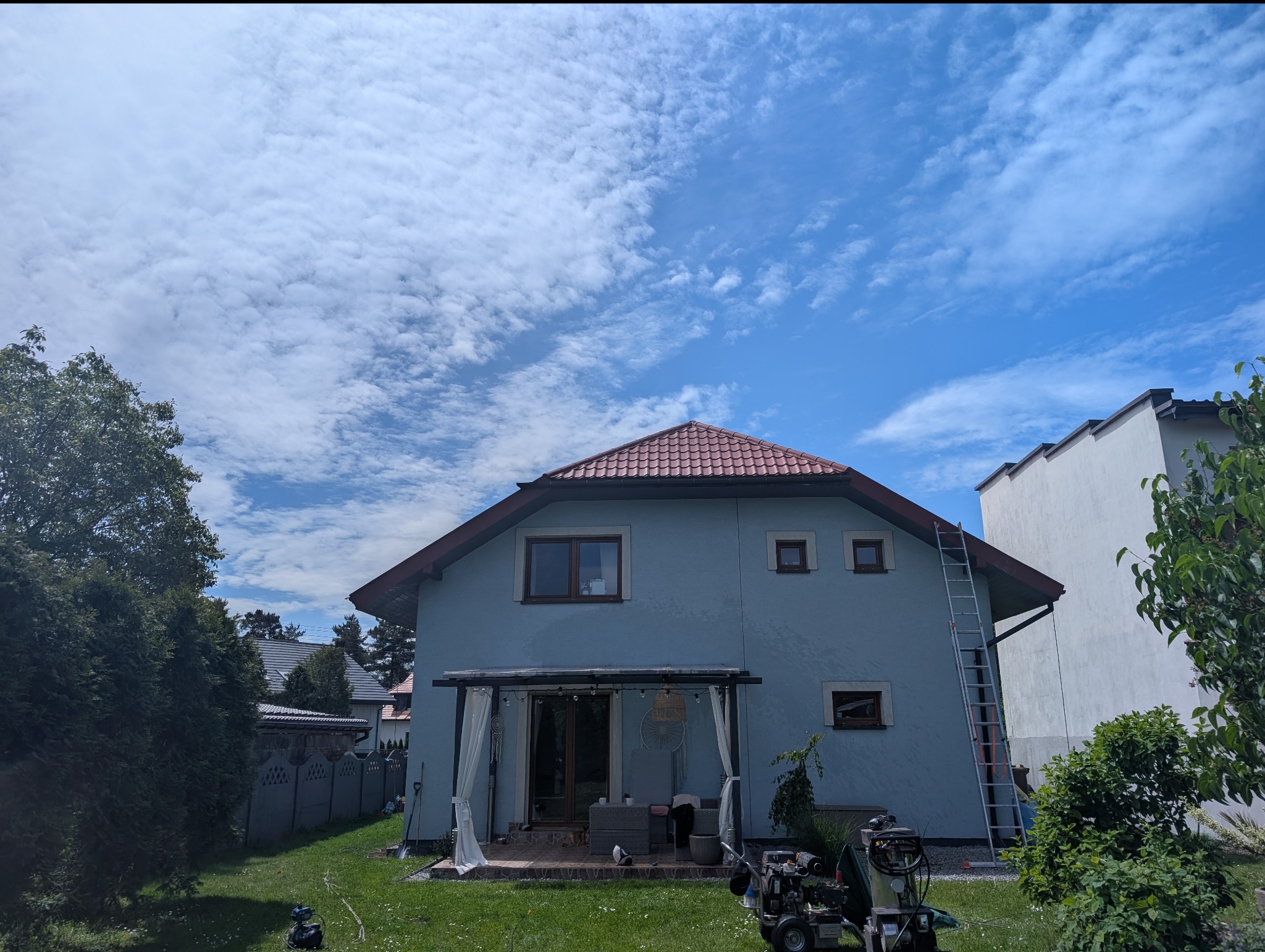 Dom z elewacją w odcieniach błękitu, z czerwoną dachówką. Przy elewacji stoi drabina i myjka ciśnieniowa. Widoczne niebo z chmurami i trawnik.