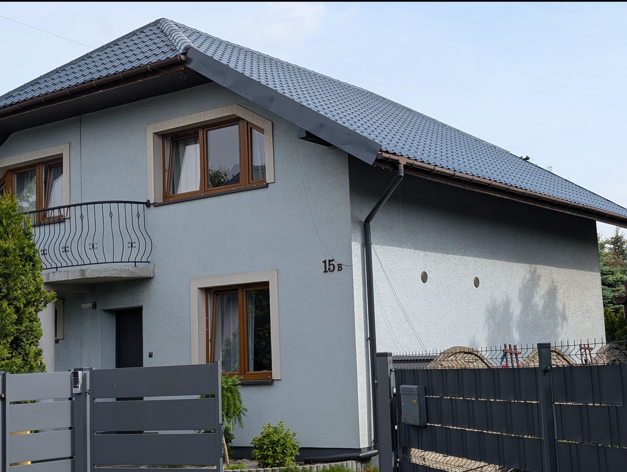 Dom jednorodzinny z szarą elewacją i ciemnym dachem z blachodachówki, widoczne rynny, okna z brązowymi ramami i balkon z ozdobną balustradą. Numer domu 15B.