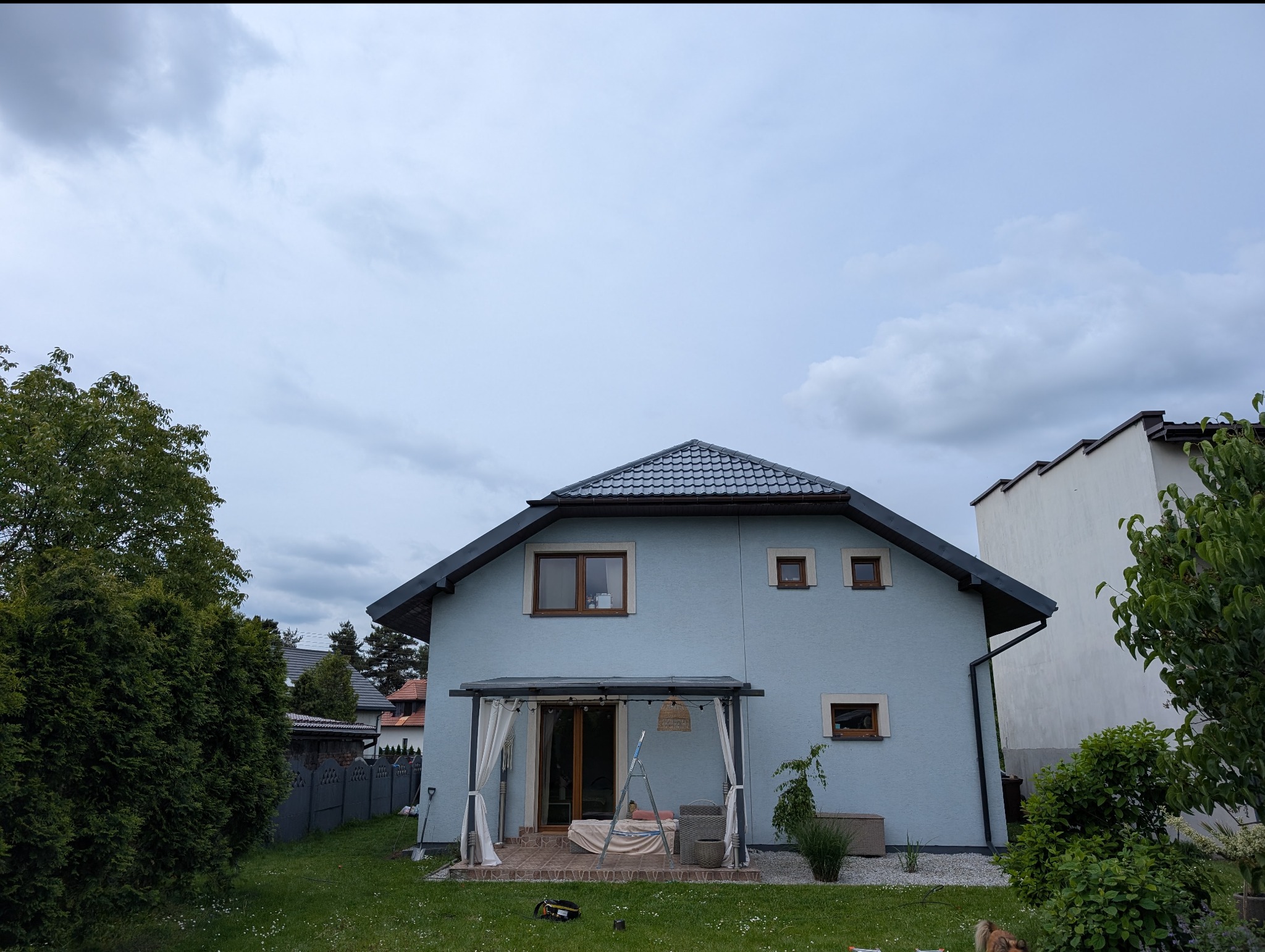 Błękitny dom jednorodzinny z szarym dachem i brązowymi ramami okiennymi, otoczony zielonym ogrodem pod pochmurnym niebem. Widoczna pergola z białymi zasłonami.