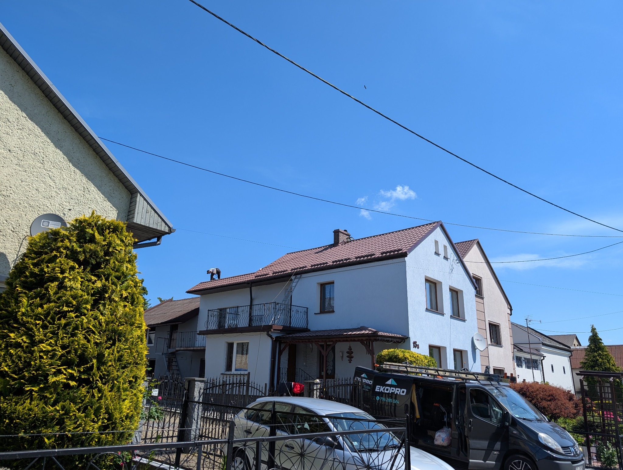 Praca na dachu: mężczyzna na dachu domu z brązową dachówką, oparty o drabinę. Na podjeździe zaparkowane auta i bus z logo firmy. Błękitne niebo w tle.