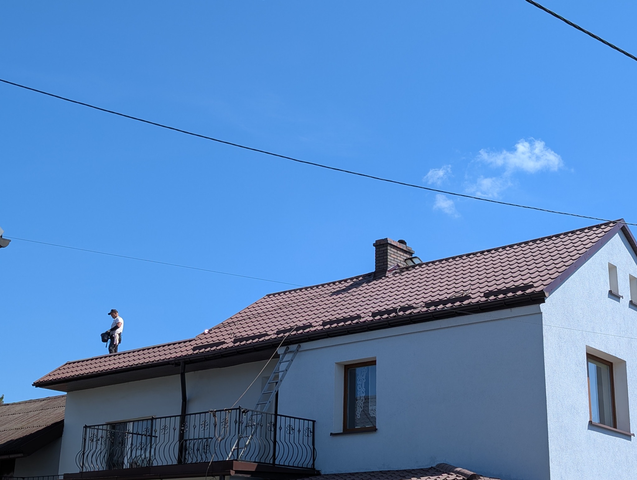 Pracownik na dachu domu z brązową dachówką, z drabiną opartą o rynnę. Błękitne niebo w tle. Widok z dołu na dwupiętrowy budynek.
