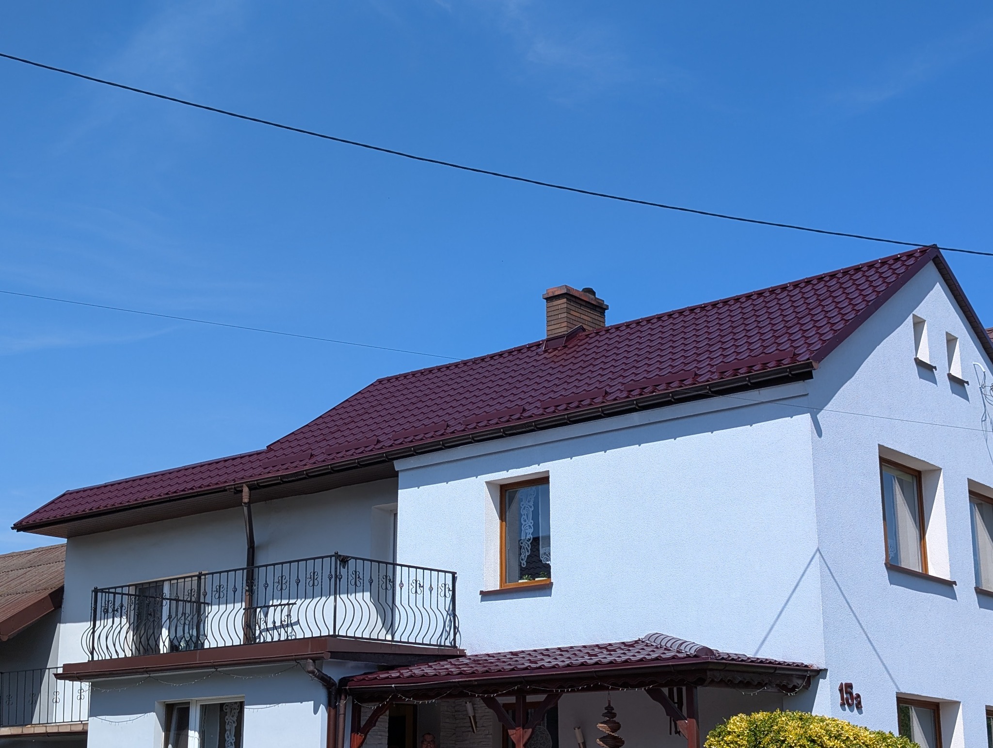Dwupiętrowy dom z ciemnoczerwonym dachem z blachodachówki i jasną elewacją, widok z dołu na tle błękitnego nieba w słoneczny dzień.