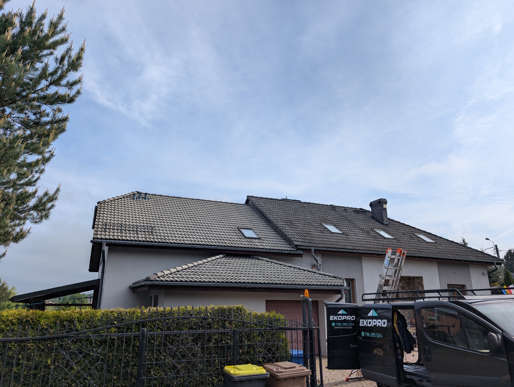 Dom z szarą dachówką i oknami dachowymi widziany z poziomu ulicy, z samochodem firmy dekarskiej na pierwszym planie i drabiną opartą o ścianę.