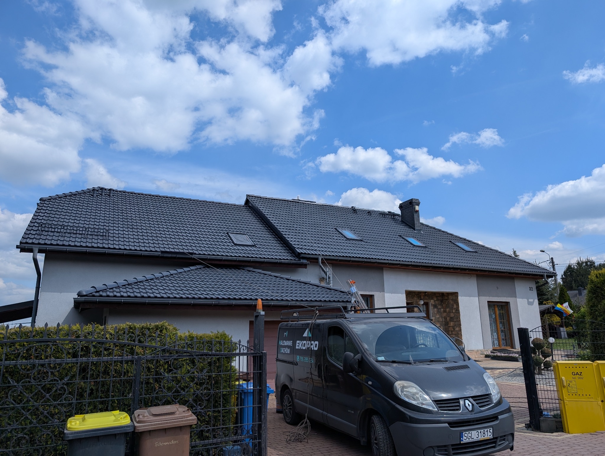 Dom z ciemną dachówką po malowaniu, na tle błękitnego nieba z chmurami. Na podjeździe bus firmy z drabiną na dachu. Widoczne kosze na śmieci i skrzynka gazowa.