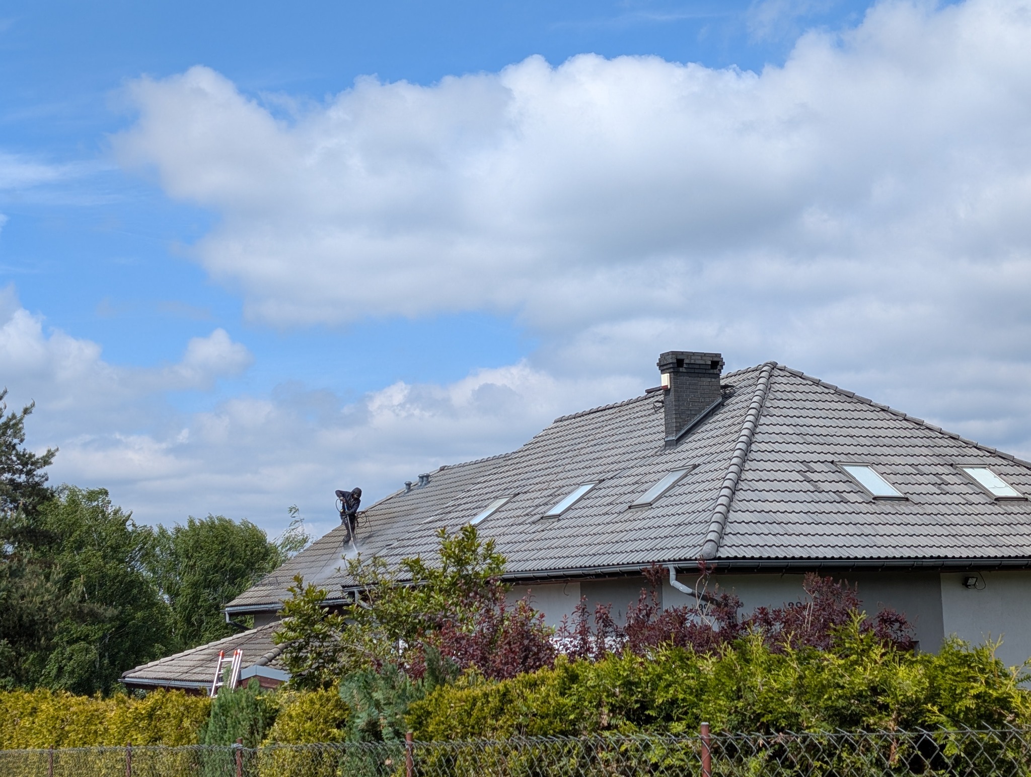 Pracownik w kombinezonie czyści dach z szarej dachówki myjką ciśnieniową. Widoczne okna dachowe, komin, zieleń w tle i fragment drabiny.