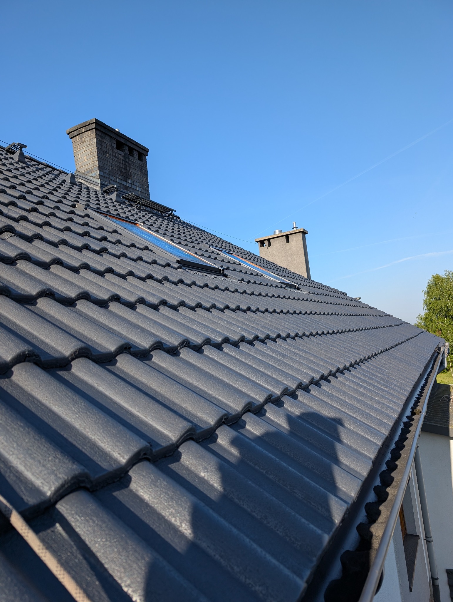Ciemnoszary dach z dachówki ceramicznej, widok z dołu pod kątem, z kominem w tle i fragmentem okna dachowego. Widoczne rynny.