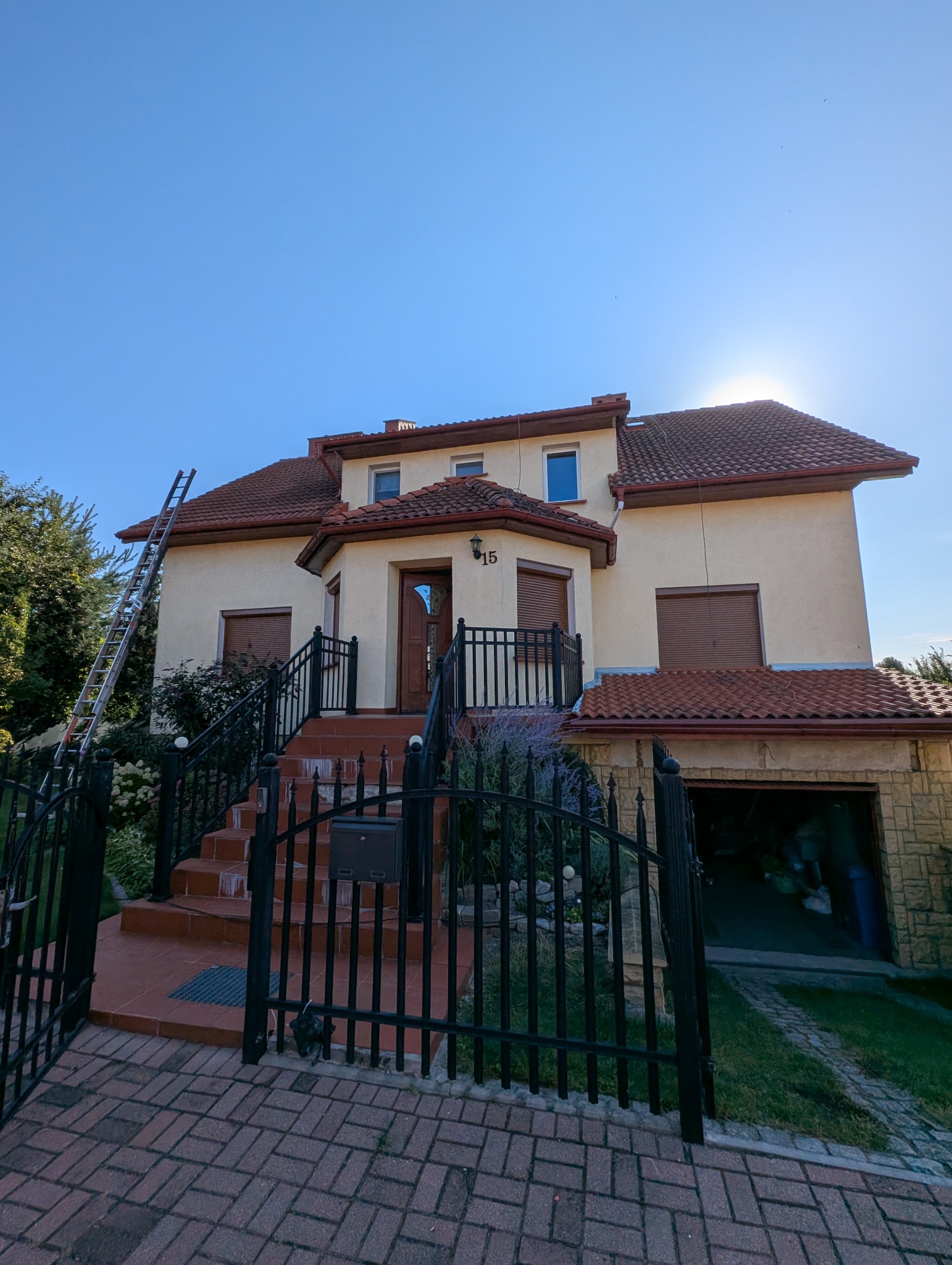 Dwupiętrowy dom z beżową elewacją i czerwoną dachówką, z rozstawioną drabiną. Widoczna kostka brukowa i czarne, metalowe ogrodzenie.