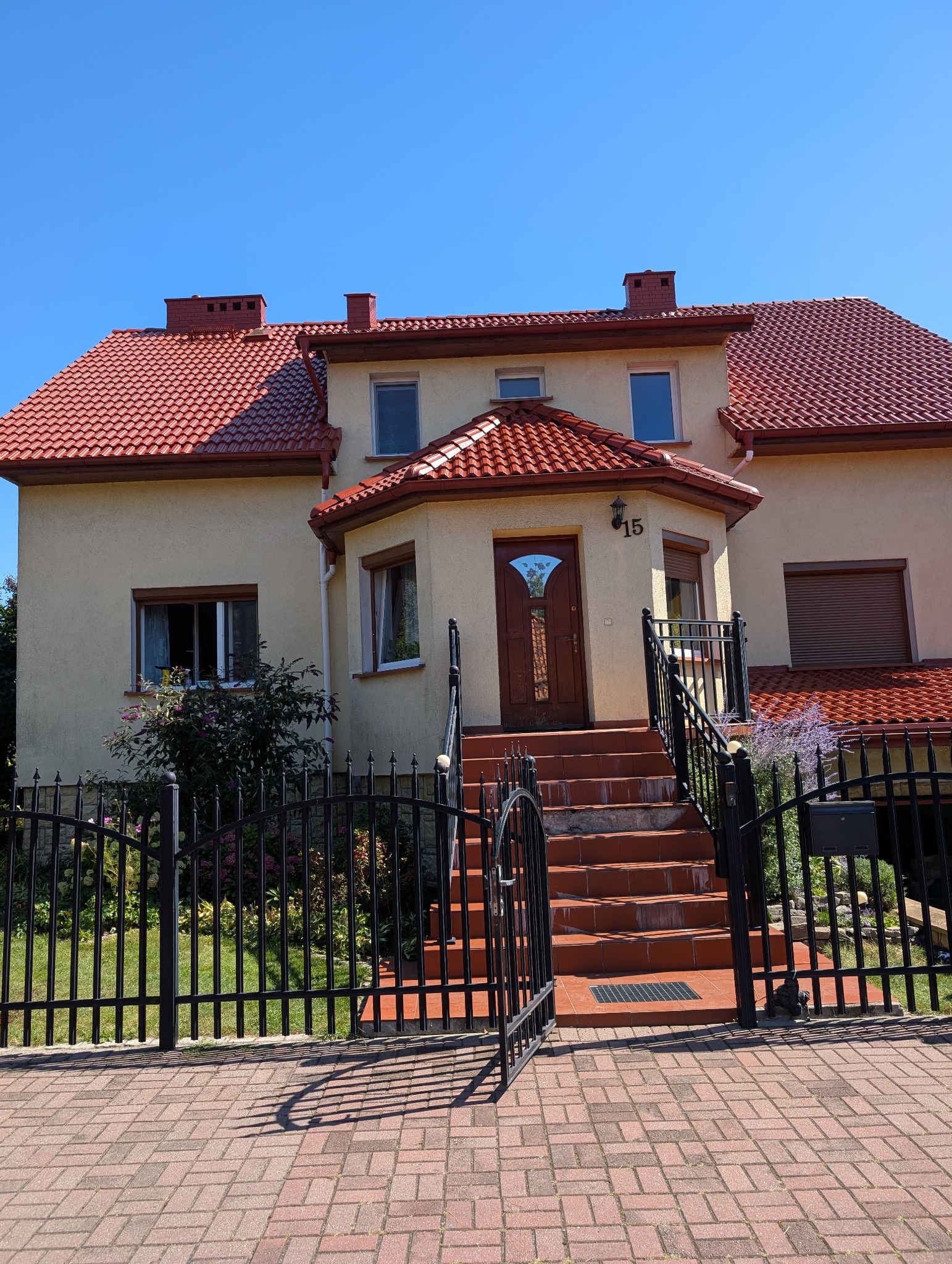 Ujęcie domu z czerwonym dachem w Pyskowicach, widoczne schody wejściowe i czarne, metalowe ogrodzenie. Dom o jasnej elewacji w słoneczny dzień.