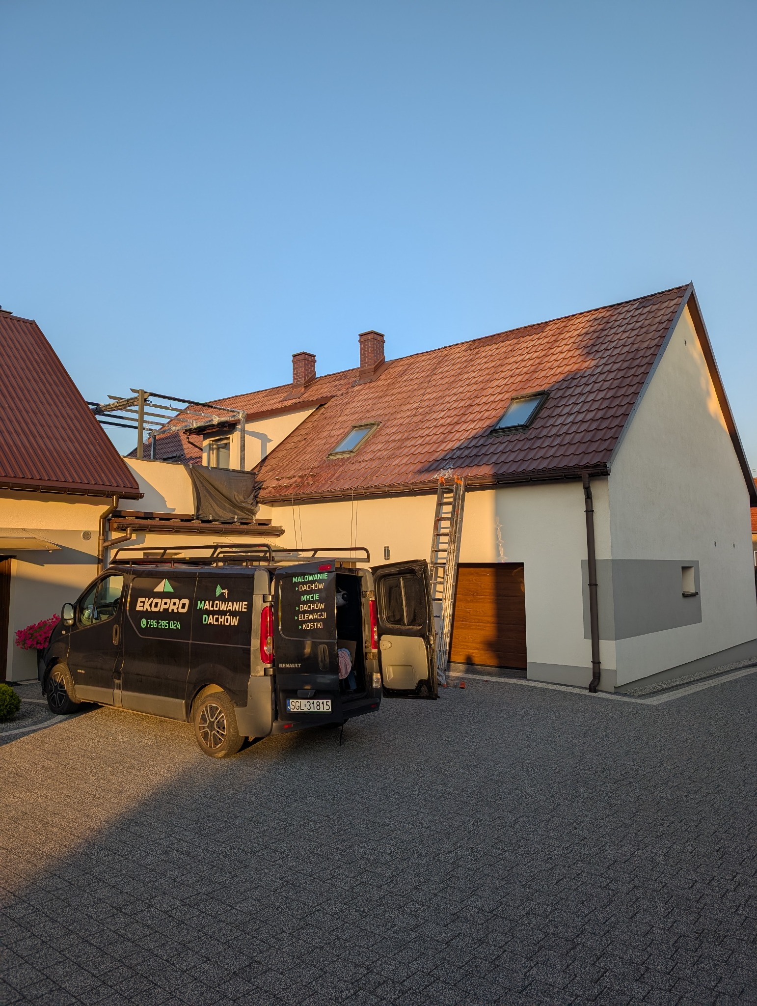 Czarny van firmy EkoPro Malowanie Dachów z otwartymi drzwiami na tle jasnego domu z brązowym dachem. Widoczna drabina i kostka brukowa.
