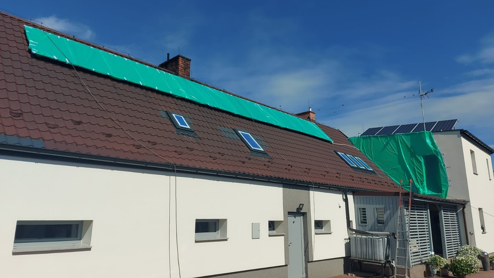 Dom z brązowym dachem krytym zieloną plandeką. Widoczne okna dachowe i panele słoneczne. Przygotowanie do malowania dachu lub elewacji.