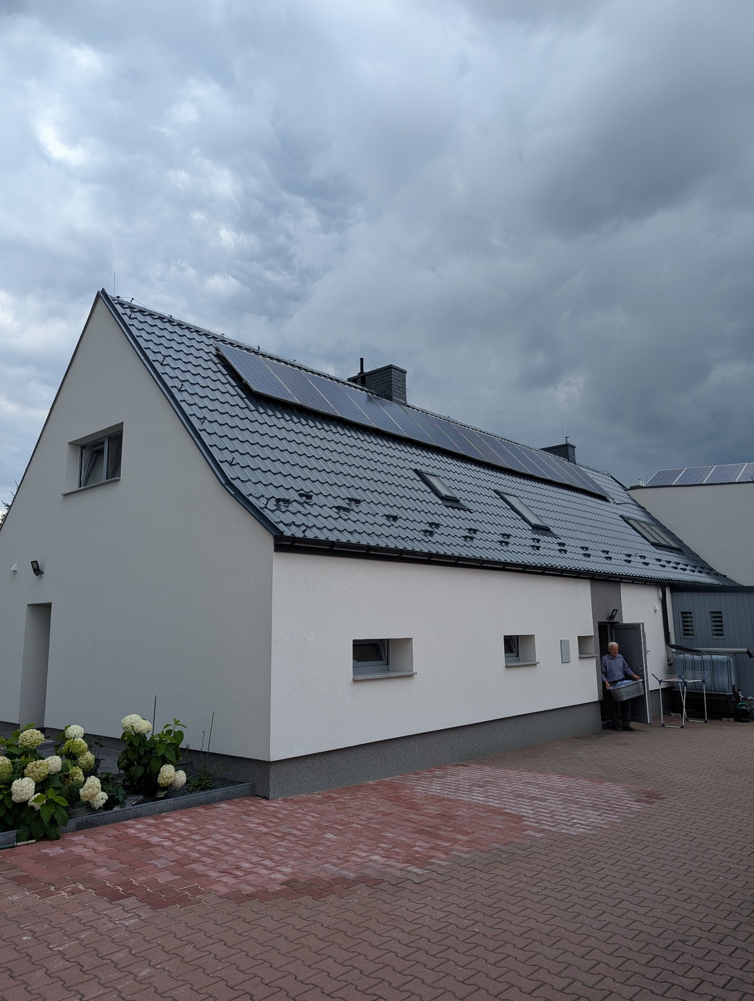 Biały dom z ciemnoszarą dachówką i panelami solarnymi. Widoczny fragment kostki brukowej i osoba w drzwiach. Pochmurne niebo w tle.