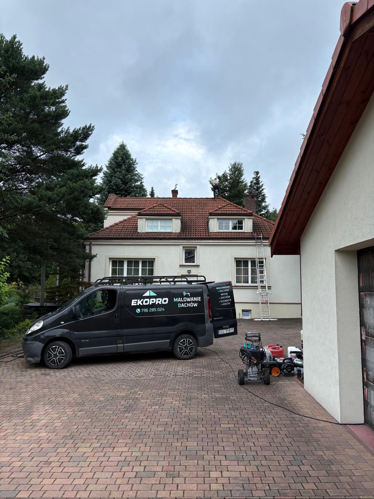 Pracownik na dachu domu z czerwonej dachówki, obok zaparkowany czarny bus firmy EkoPro Malowanie Dachów i sprzęt do mycia pod ciśnieniem. Pochmurne niebo.