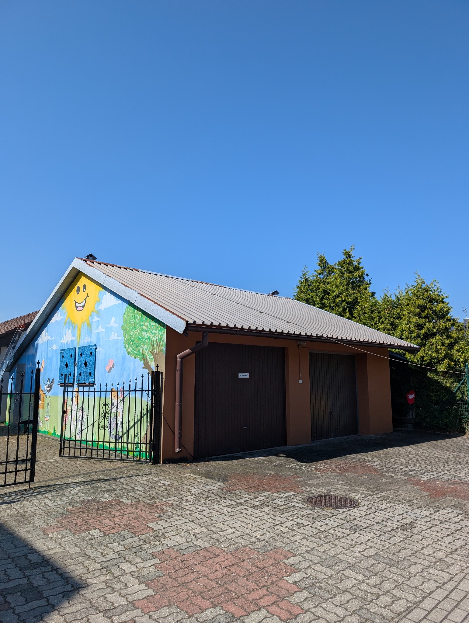 Kolorowy garaż z muralem słońca i drzew, metalowy dach, brązowe bramy garażowe, kostka brukowa i czarne ogrodzenie. Błękitne niebo w tle.