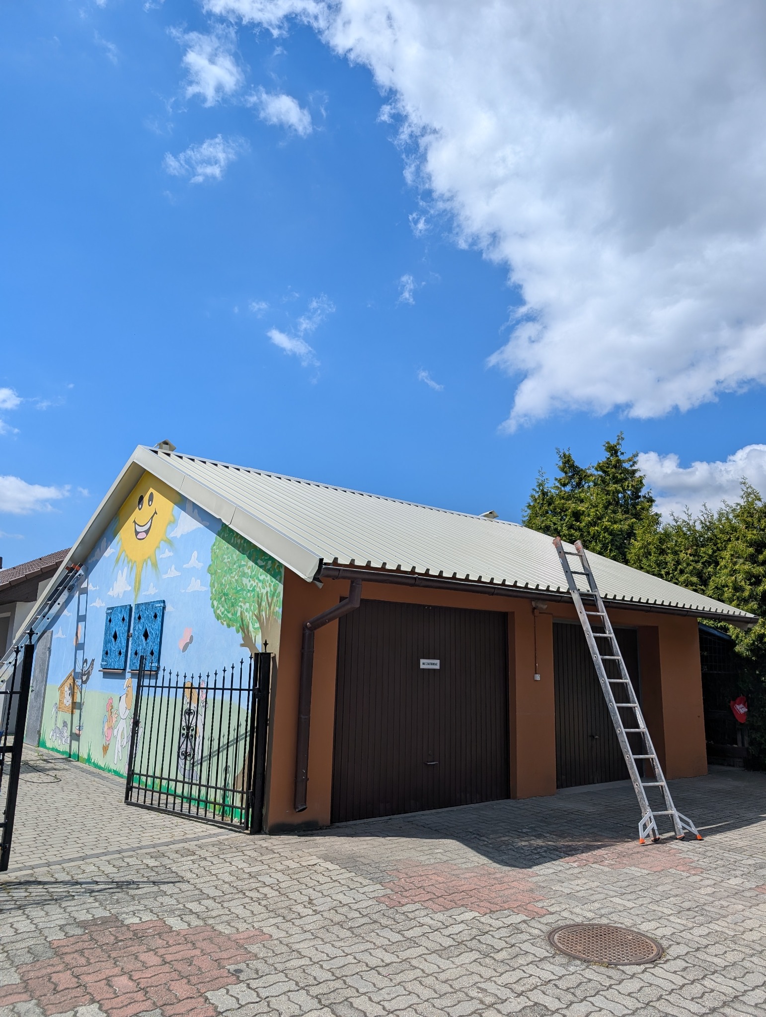 Dwukondygnacyjny budynek z pomalowaną ścianą w bajkowe motywy, metalowym dachem i dwoma bramami garażowymi. Przy dachu oparta drabina.