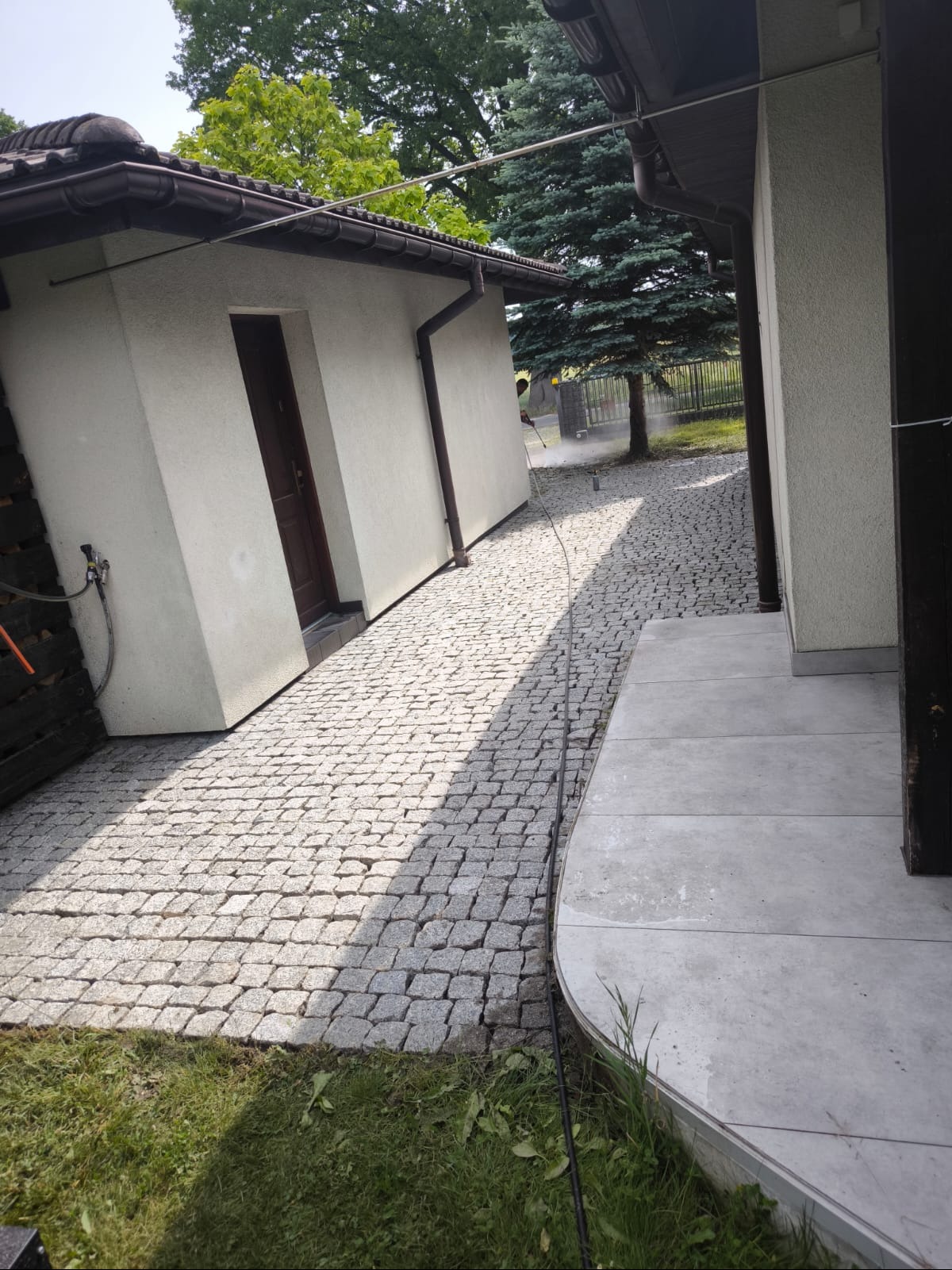 Czyszczenie kostki brukowej na posesji prywatnej za pomocą myjki ciśnieniowej. Widoczna osoba w trakcie pracy, budynek gospodarczy z brązowymi elementami.