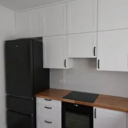 Nowoczesna kuchnia z białymi frontami szafek, drewnianym blatem i czarną lodówką oraz piekarnikiem.