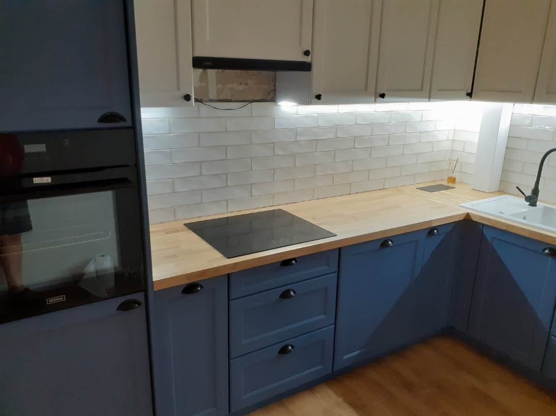 Niebiesko-biała kuchnia z drewnianym blatem, płytą indukcyjną i białymi płytkami na ścianie. Widoczny fragment piekarnika w zabudowie i czarne uchwyty szafek.