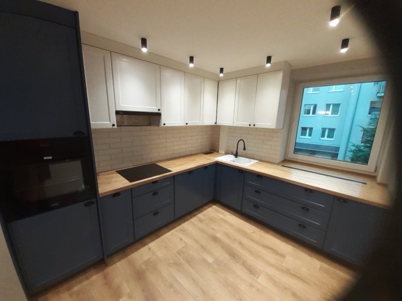 Dwukolorowa kuchnia w kształcie litery L z białymi szafkami wiszącymi i niebieskimi dolnymi, drewnianym blatem, białymi płytkami ściennymi imitującymi cegłę oraz widokiem na budynek zza okna.