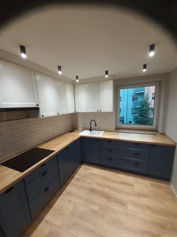 Nowoczesna kuchnia w kształcie litery L z białymi szafkami górnymi i granatowymi dolnymi, drewnianym blatem, białym zlewem i czarną armaturą, oświetlona punktowymi lampami sufitowymi, z widokiem...