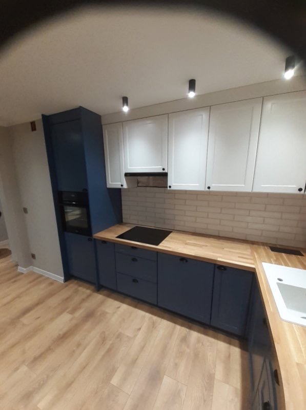 Nowoczesna kuchnia z białymi szafkami wiszącymi i granatowymi szafkami dolnymi, drewniany blat, płytki imitujące cegłę na ścianie, oświetlenie punktowe.