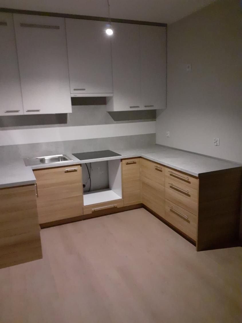 Jasna kuchnia z drewnianymi frontami szafek dolnych i białymi górnymi, szary blat, płyta indukcyjna i zlewozmywak w rogu, widoczne kable i rury pod zlewem.