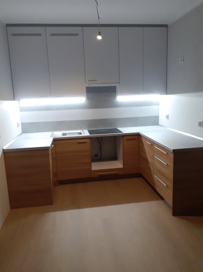 Jasna kuchnia z drewnianymi szafkami dolnymi i szarymi górnymi, podświetlona listwami LED pod szafkami wiszącymi, widok na narożnik blatu z wbudowanym zlewem i płytą grzewczą.