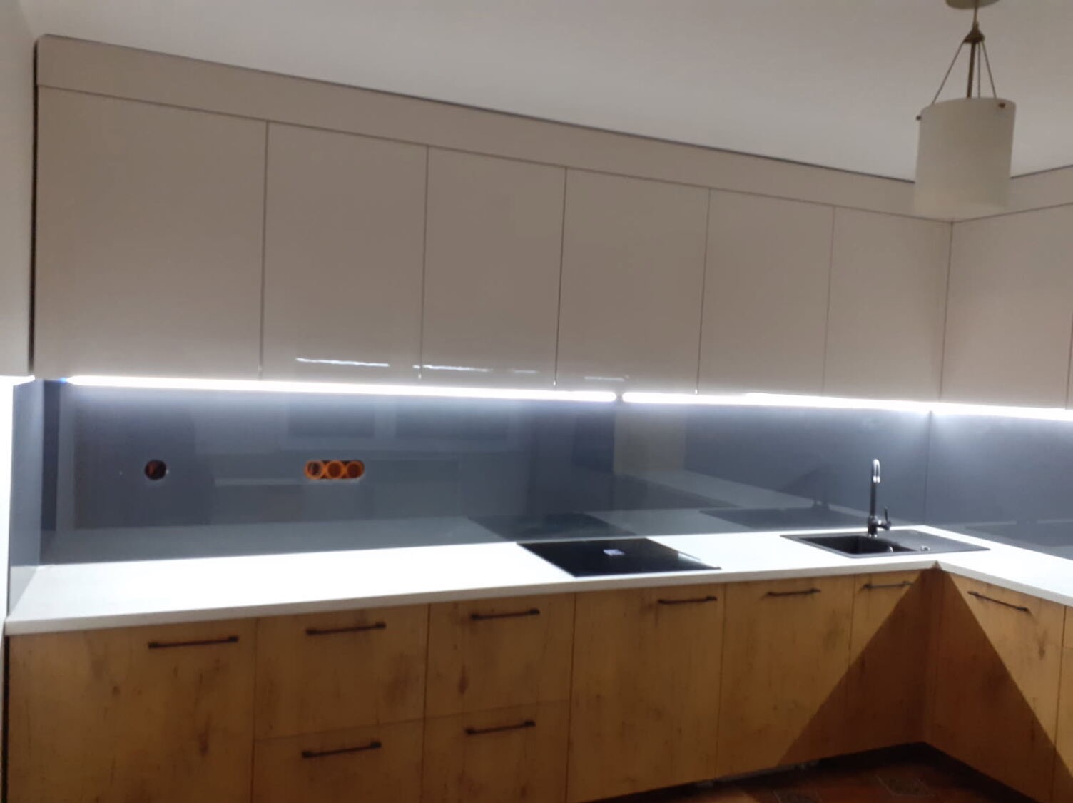 Nowoczesna kuchnia z drewnopodobnymi szafkami dolnymi, białym blatem i szarym panelem szklanym podświetlonym LED, widoczne gniazdka elektryczne.