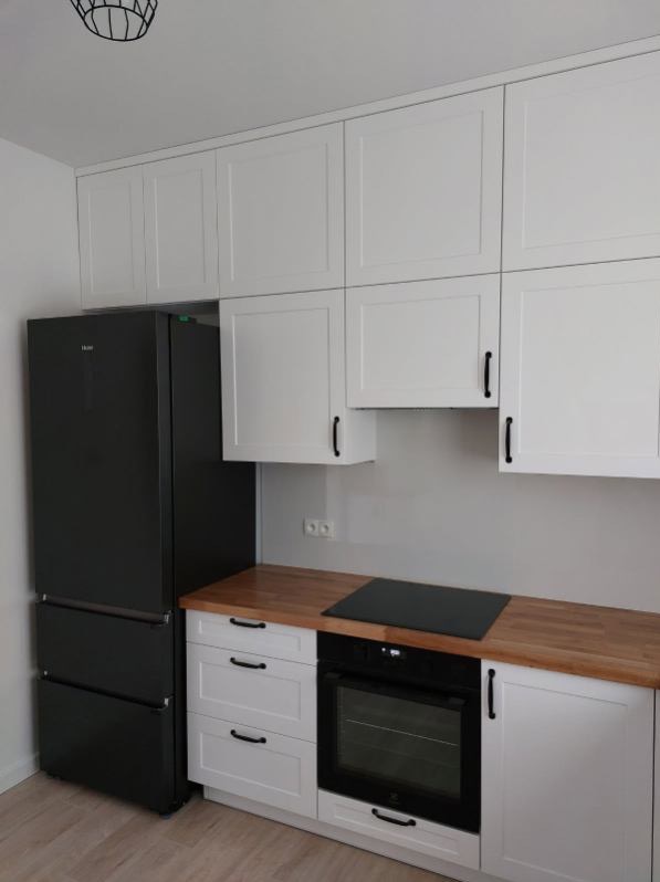 Nowoczesna kuchnia z białymi frontami szafek, drewnianym blatem i czarną lodówką oraz piekarnikiem.