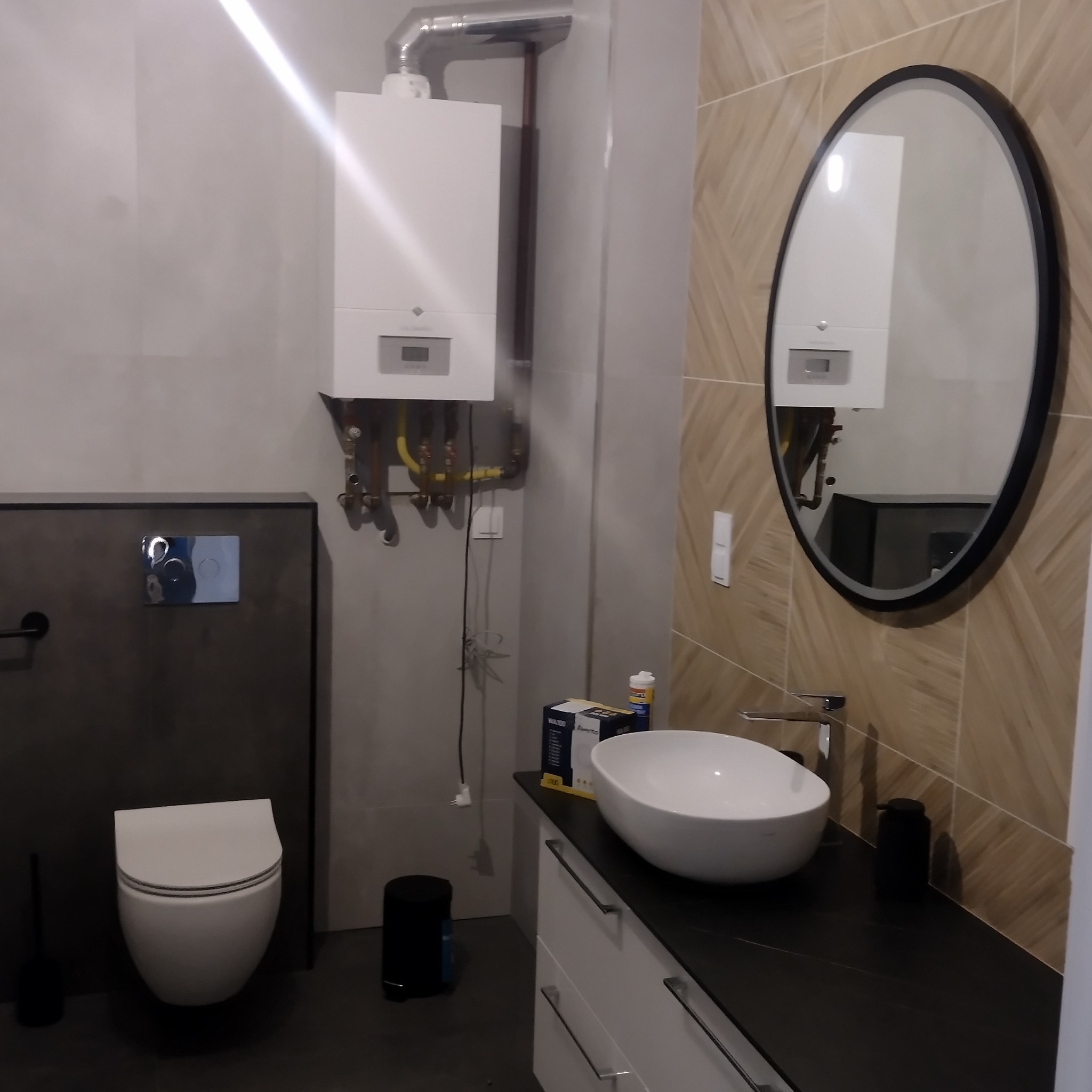 Nowoczesna łazienka z owalnym lustrem, wiszącą toaletą, białą umywalką nablatową i grzejnikiem gazowym na ścianie.