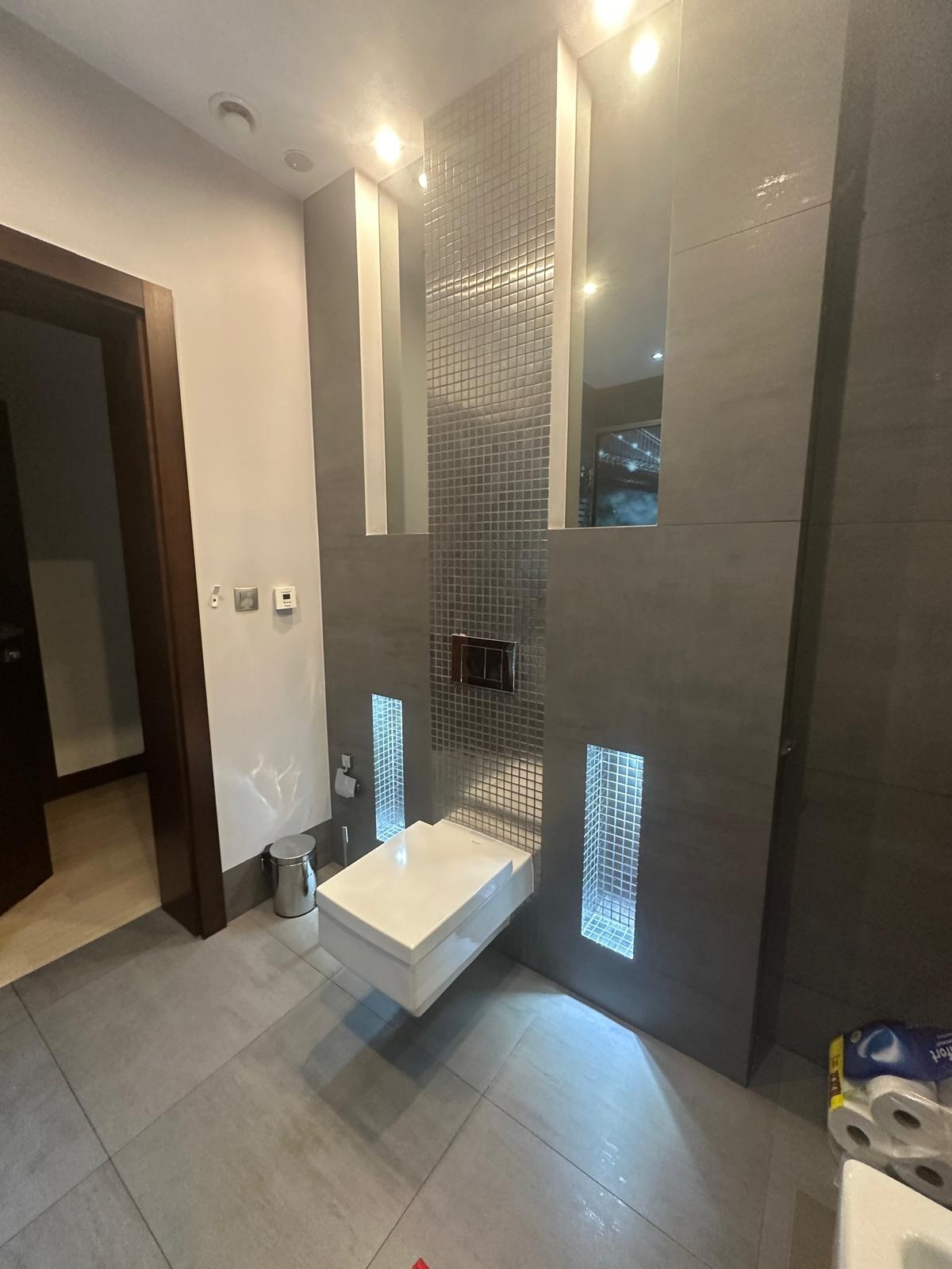 Nowoczesna łazienka z podwieszaną toaletą, szarą mozaiką i podświetlanymi niszami, widoczne rolki papieru toaletowego w rogu