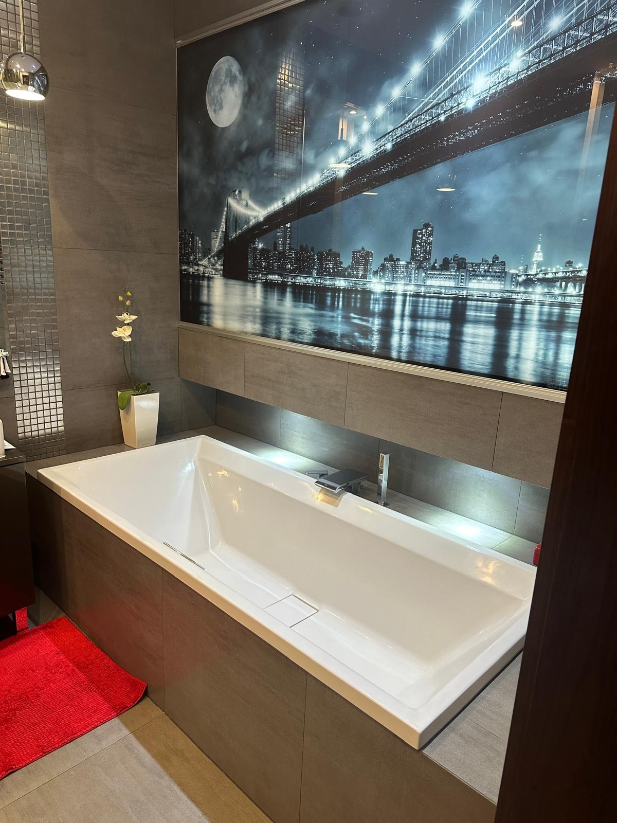 Nowoczesna łazienka z białą wanną, szarą glazurą i fototapetą z nocnym widokiem na most i miasto, podświetlona listwą LED.