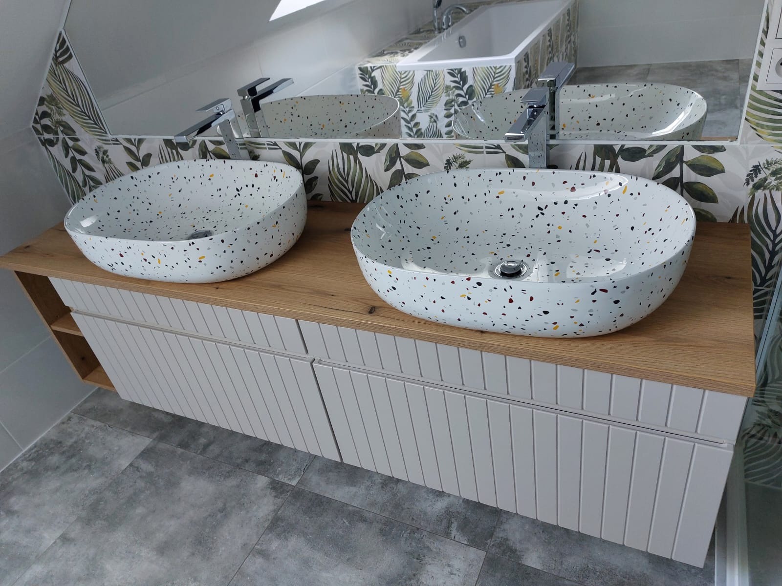 Dwie umywalki w stylu lastryko na drewnianym blacie z szafką w łazience w Radziemicach, z widocznym fragmentem wanny obłożonej płytkami z motywem roślinnym.