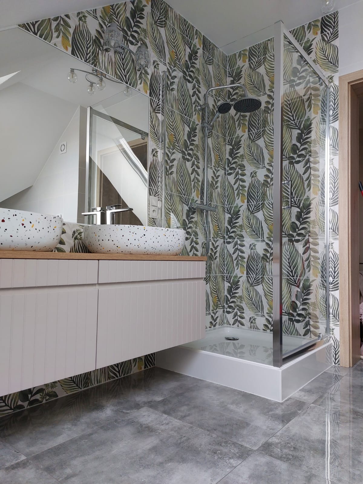 Nowoczesna łazienka z umywalkami nablatowymi w stylu terrazzo, szafką w kolorze pudrowego różu i kabiną prysznicową, ściany wyłożone płytkami z motywem tropikalnych liści.