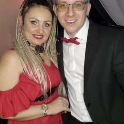 Zespół Muzyczny DEXTER - Portret kobiety w czerwonym kombinezonie i mężczyzny w czarnym garniturze z czerwoną muszką, pozujących na tle białych zasłon i oświetlenia scenicznego.