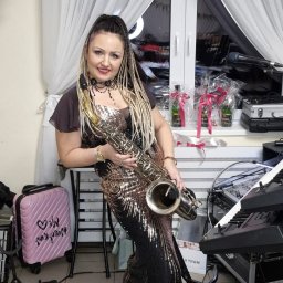 Zespół Muzyczny DEXTER - Kobieta w cekinowej sukni pozuje z saksofonem na tle okna z białymi firankami i sprzętu muzycznego.