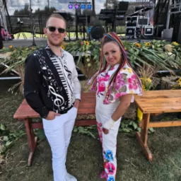 Uśmiechnięty duet muzyczny na tle sceny plenerowej: mężczyzna w koszuli z motywem klawiatury, kobieta w kwiecistym stroju i kolorowych warkoczykach. Scena udekorowana słonecznikami.