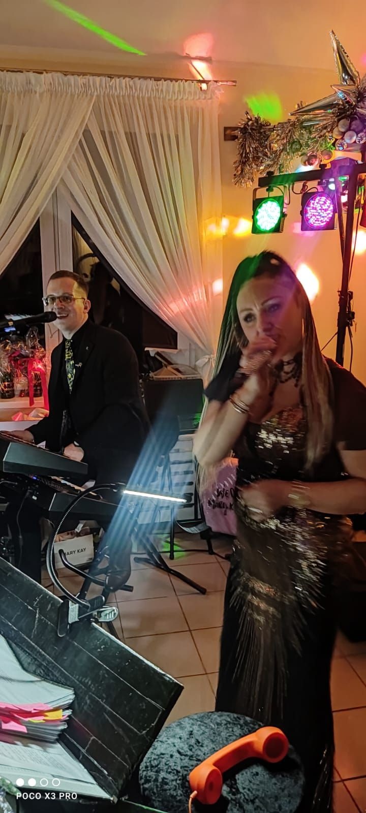 Kobieta w cekinowej sukience śpiewa do mikrofonu, obok mężczyzna gra na keyboardzie, za nimi dekoracje i kolorowe oświetlenie sceniczne.