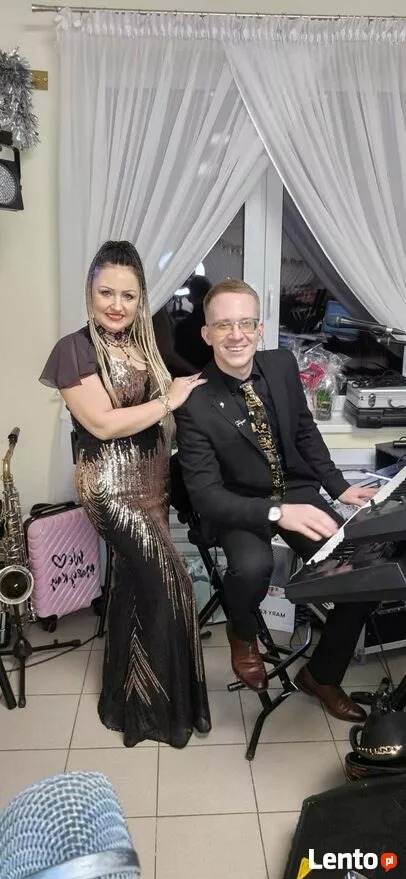 Duet muzyczny: wokalistka w złoto-czarnej sukni i pianista w czarnym garniturze, obok saksofon i różowa walizka, występ na tle okna z białymi firankami i świąteczną dekoracją.