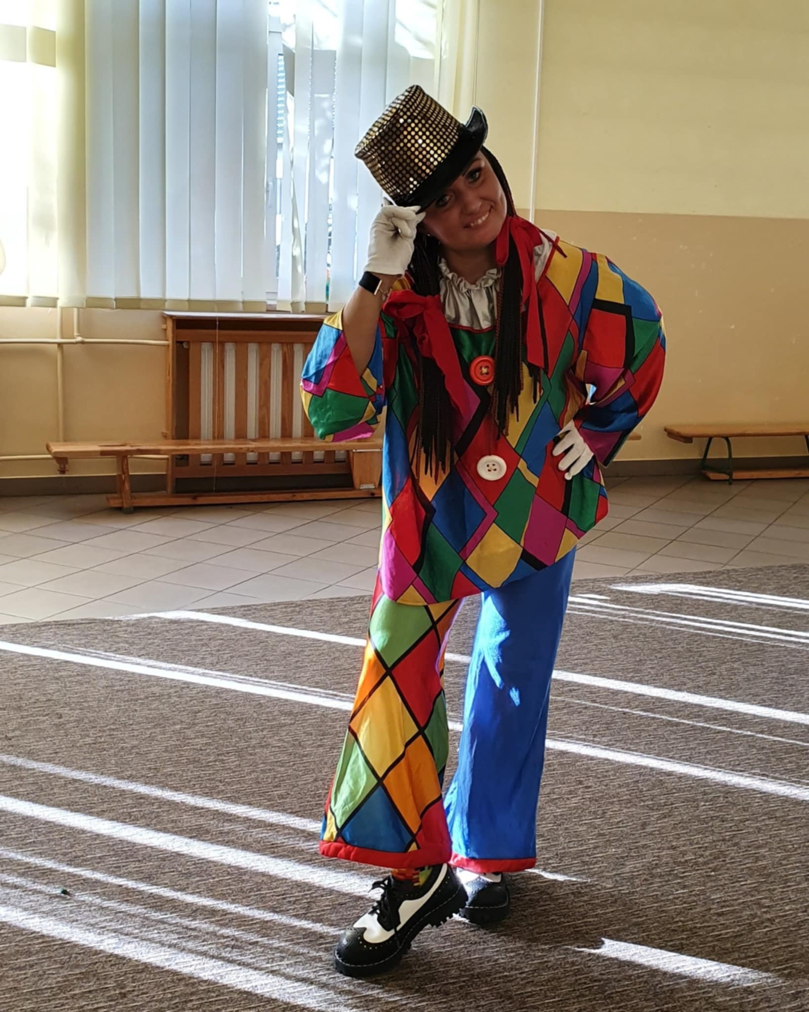 Uśmiechnięta kobieta w kostiumie klauna z kolorowymi rombami, złotym cylindrem i białych rękawiczkach, pozująca w jasnym pomieszczeniu z promieniami słońca.