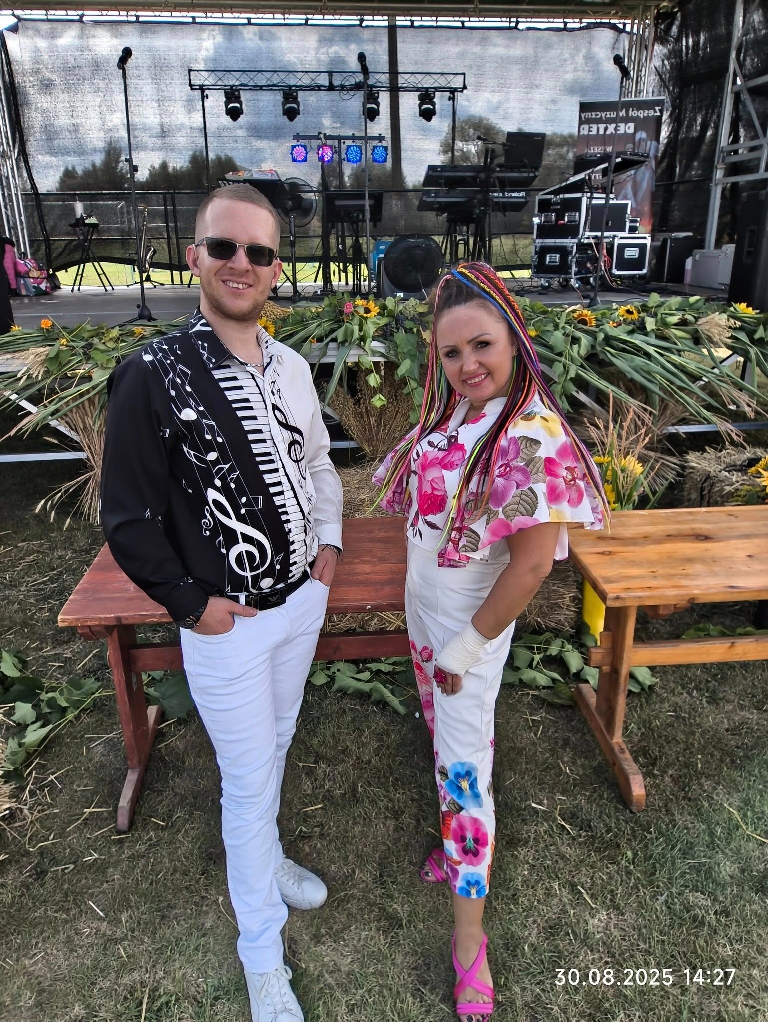 Uśmiechnięty duet muzyczny na tle sceny plenerowej: mężczyzna w koszuli z motywem klawiatury, kobieta w kwiecistym stroju i kolorowych warkoczykach. Scena udekorowana słonecznikami.