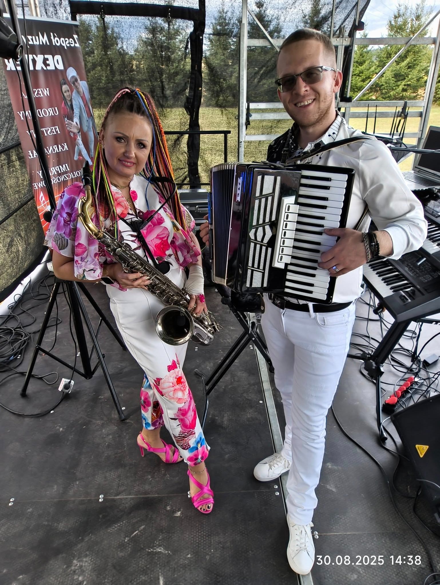 Duet muzyczny z saksofonem i akordeonem w Długołęce, na scenie plenerowej. Kobieta w kwiecistym stroju, mężczyzna w bieli. W tle baner reklamowy.