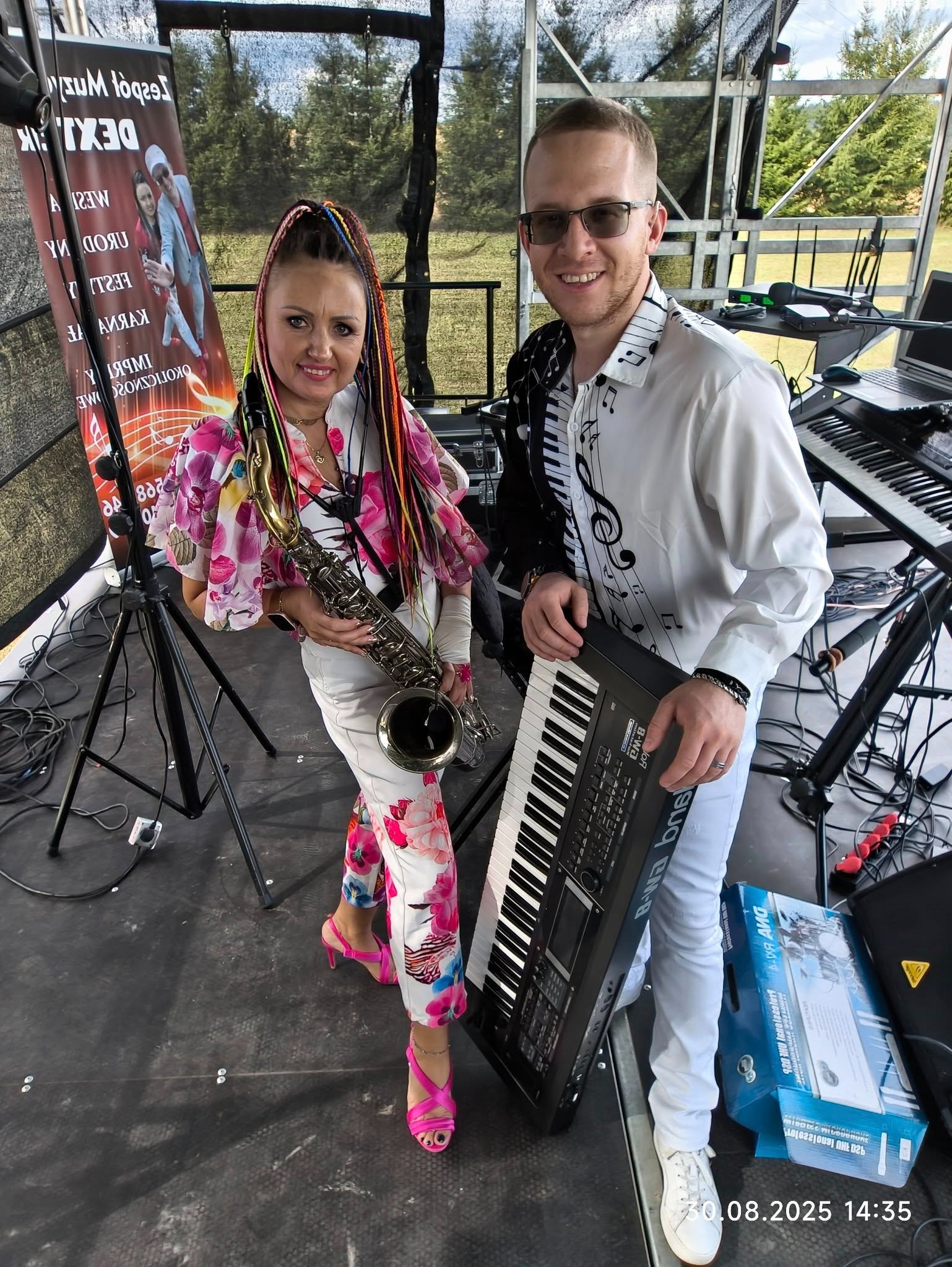 Duet muzyczny: kobieta z saksofonem i mężczyzna z keyboardem w stylizowanych strojach na scenie, z plakatem zespołu w tle. Scena plenerowa.