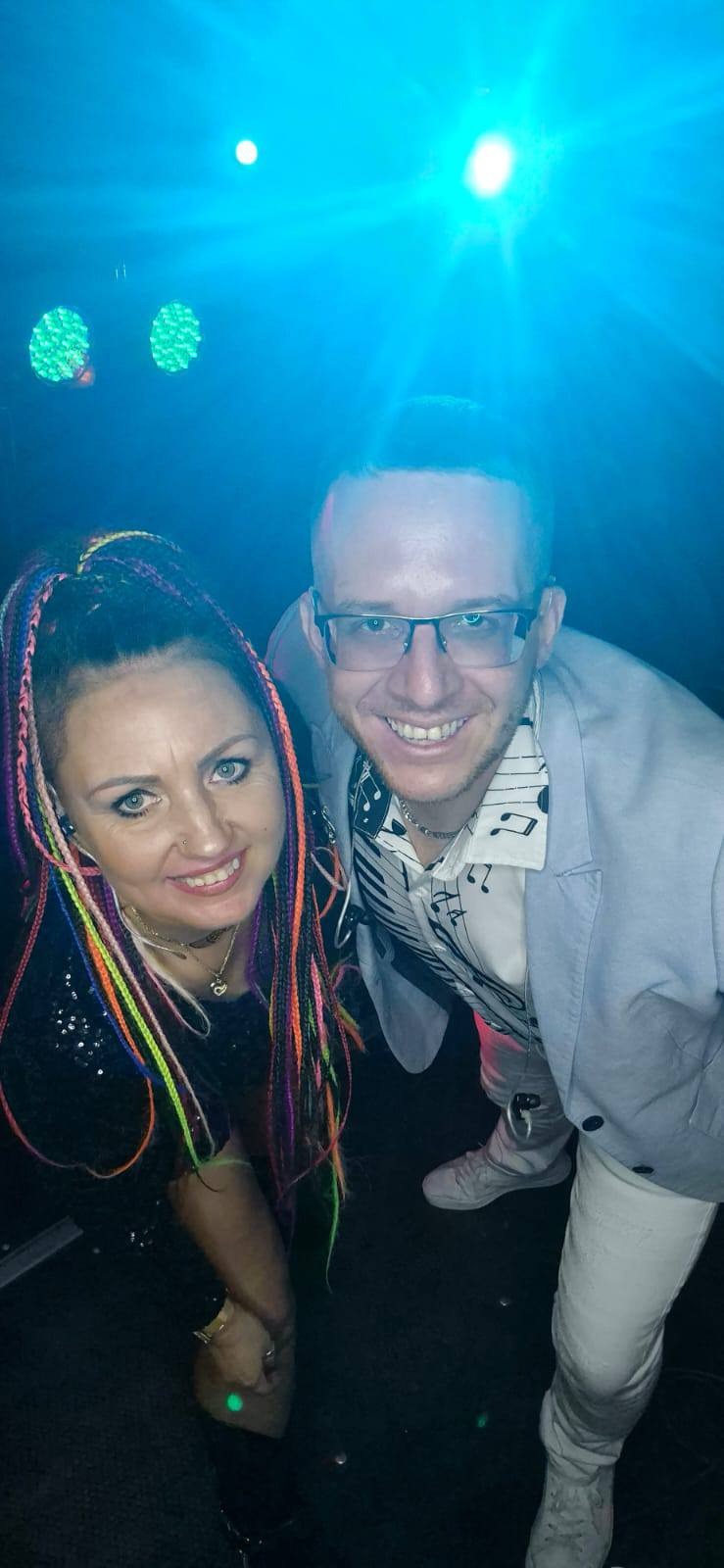 Uśmiechnięty duet muzyczny w blasku niebieskich świateł scenicznych: kobieta z kolorowymi warkoczykami i mężczyzna w marynarce i koszuli z motywem klawiatury.