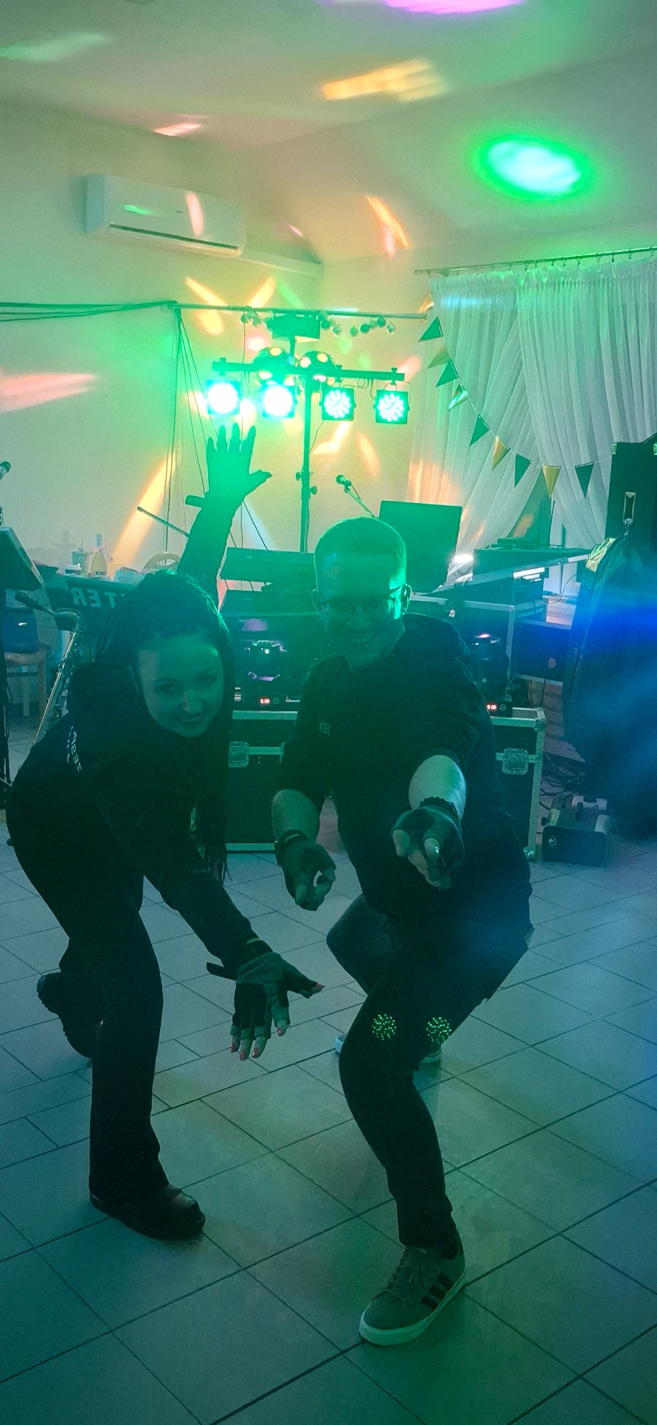 Dwoje tancerzy w czarnych strojach, oświetlonych zielonym światłem scenicznym, prezentuje dynamiczne pozy na parkiecie podczas występu. W tle sprzęt muzyczny i dekoracje.